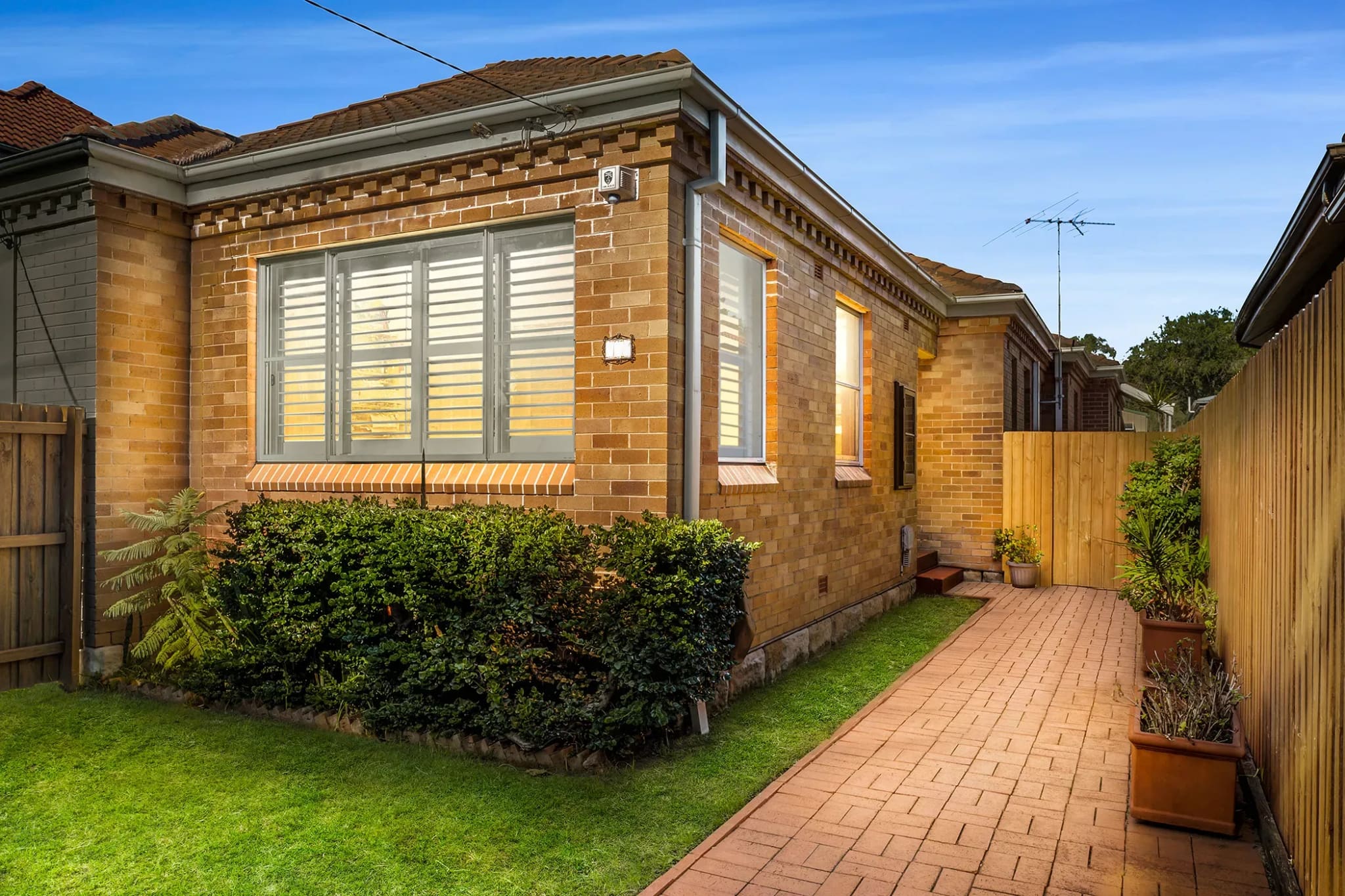 Blond-brick bungalow with shuttered windows, a clipped hedge and a brick side path at dusk, Fairlight