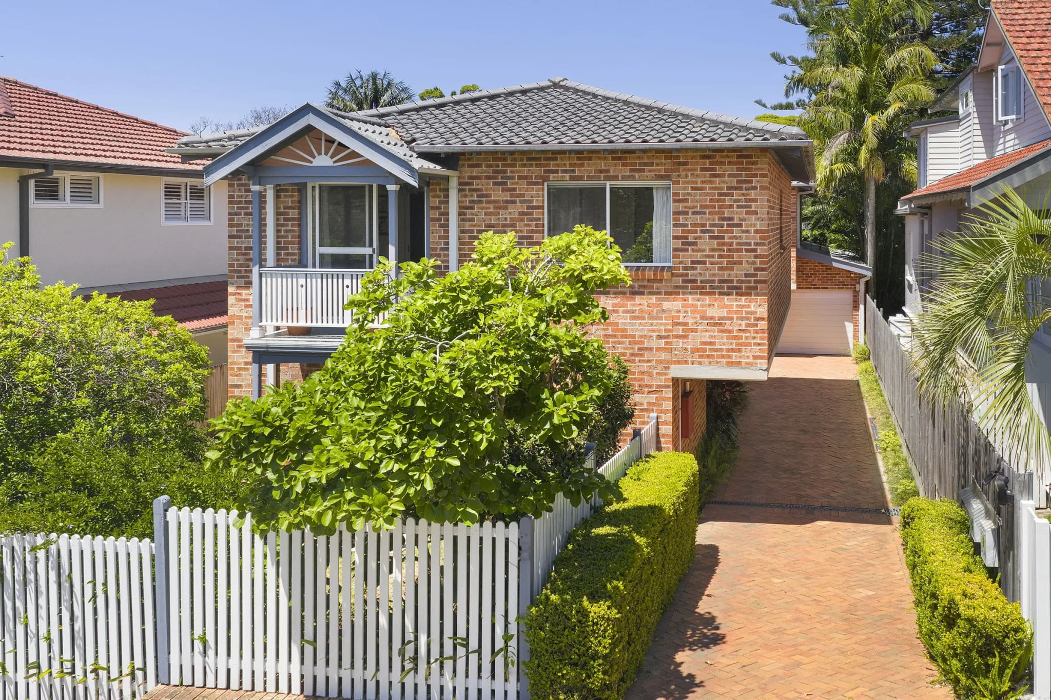 Two-storey brick townhouse with a front balcony, behind a white picket fence and a brick driveway, Fairlight