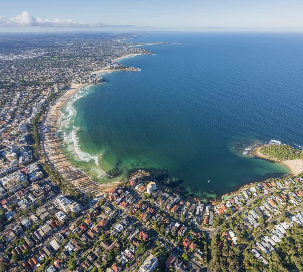 An aerial view over Manly looking north along the Northern Beaches coastline, with surf breaking the length of the beach, a rocky headland and sheltered bay in the foreground, and the coast running away to the horizon.